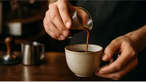 Hands of a barista gently pouring a rich espresso shot into a ceramic cup. The café background is softly blurred, with visible coffee tools like a tamper and milk pitcher. Warm lighting, dark wood counter, artisanal feel. Emphasis on process and craft.